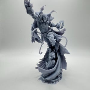 May include: A gray resin figurine depicting a muscular, masked character in a dynamic leaping pose with an outstretched arm, wearing a flowing cape and chains. Below the character, a smaller winged figure is partially enveloped by the cape.
