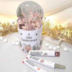 May include: A cylindrical advent calendar with a pop-up village scene. The calendar is white with the text "24 Kleine Zaubermomente". Three rolled paper treats are in front of the calendar, each with German text and a small illustration.