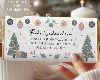 May include: A festive chocolate bar wrapper with the text "Frohe Weihnachten" and a holiday message. The design includes Christmas trees, ornaments, and stars in shades of green, blue, and gold. The wrapper is held by a person.
