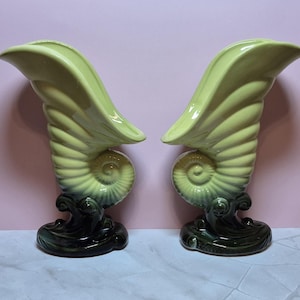 May include: Pair of ceramic vases in a seashell design. The vases are light green with a spiral pattern and dark green wave-like bases. The vases have a curved, wing-like top.