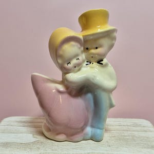 May include: A ceramic figurine of a couple embracing, with the woman in a pink dress and the man in a blue suit and yellow top hat. The figurine is on a white wooden stand, with a pink background.