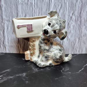 May include: A ceramic planter shaped like a mailbox with a seated dog figurine. The mailbox is cream-colored with a purple flag, and the dog has black and gray spots. The base is brown with a textured design.