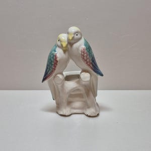 May include: A ceramic figurine featuring two parakeets perched on a white tree stump. The birds have white bodies, yellow heads, and wings with blue and pink scale-like patterns. The base is a hollow white structure with a central opening.