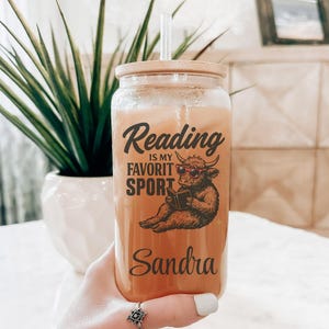 May include: A clear glass tumbler with a wooden lid and straw, filled with a light orange beverage. The tumbler features the text "Reading is my favorite sport" and a cartoon bull reading a book. The name "Sandra" is printed below.