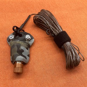 May include: A camouflage-patterned housing unit with two silver-colored metal bolts and a copper-toned cylindrical knob, connected by a black wire to a coiled length of brown cable secured with a black hook-and-loop fastener strap.