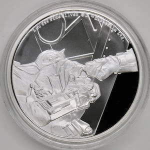 Puede incluir: Una moneda de plata con una imagen detallada de un personaje de la franquicia Star Wars, conocido como "The Child" o "Baby Yoda", que extiende la mano hacia otra. La moneda está inscrita con "1 oz 999 Fine Silver" y "Lucasfilm Ltd."