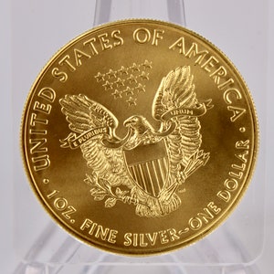May include: A gold-colored United States of America one ounce silver dollar coin with an image of an eagle with outstretched wings and a shield on its chest. The coin has the words "United States of America" and "One Dollar" inscribed around the edge. The coin also has the words "E Pluribus Unum" and "1 oz. Fine Silver" inscribed on it.