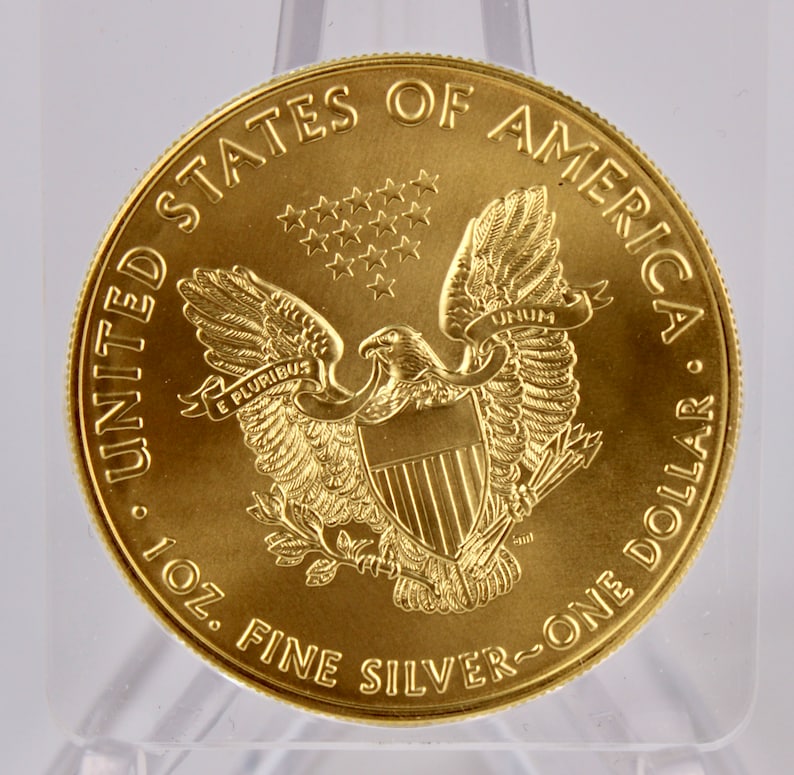 May include: A gold-colored United States of America one ounce silver dollar coin with an image of an eagle with outstretched wings and a shield on its chest. The coin has the words "United States of America" and "One Dollar" inscribed around the edge. The coin also has the words "E Pluribus Unum" and "1 oz. Fine Silver" inscribed on it.