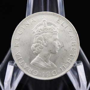 May include: A silver coin featuring a profile of a woman wearing a crown, surrounded by text. The coin is displayed on a clear plastic stand against a black background. The coin's details are finely rendered.