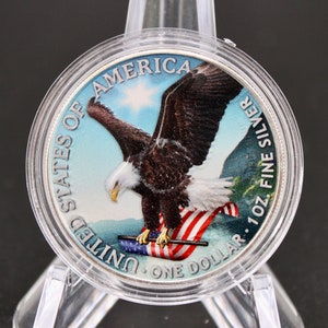 May include: A silver one dollar coin featuring a bald eagle with outstretched wings, holding a US flag. The coin is inscribed "United States of America, One Dollar, 1 oz. Fine Silver".