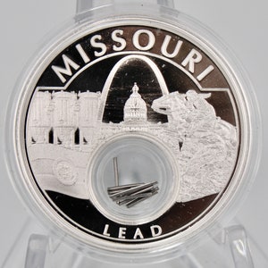 May include: A silver coin with the word "MISSOURI" around the edge and a depiction of the Gateway Arch, the state capitol building, and a horse and rider. The coin also has the word "LEAD" on the bottom.