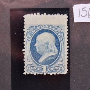 May include: A vintage blue one-cent U.S. postage stamp featuring a profile of Benjamin Franklin. The stamp is framed with ornate scrollwork and the words "U.S. POSTAGE" and "ONE CENT". The stamp is mounted on a black background.