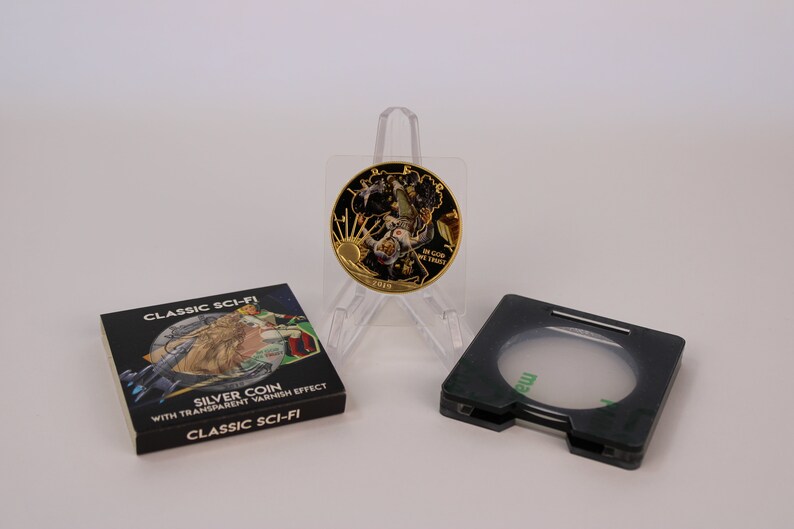 May include: A gold-colored coin with a black and gold design featuring a space scene. The coin is on a clear plastic stand. The coin is packaged in a black box with the text "CLASSIC SCI-FI SILVER COIN WITH TRANSPARENT VARNISH EFFECT CLASSIC SCI-FI" printed on it.