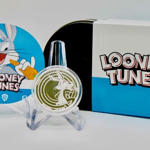 May include: A silver coin featuring Bugs Bunny from Looney Tunes. The coin is on a clear display stand. The coin is packaged in a blue and white box with the Looney Tunes logo.
