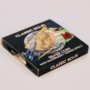 May include: A black box with a silver coin inside. The box has a design of a woman in a dress and a spaceship. The text on the box reads "CLASSIC SCI-FI SILVER COIN WITH TRANSPARENT VARNISH EFFECT CLASSIC SCI-FI".
