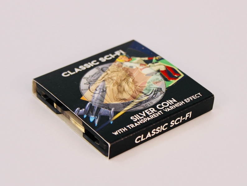 May include: A black box with a silver coin inside. The box has a design of a woman in a dress and a spaceship. The text on the box reads "CLASSIC SCI-FI SILVER COIN WITH TRANSPARENT VARNISH EFFECT CLASSIC SCI-FI".