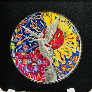 May include: A silver coin with a colorful design featuring a winged figure and abstract patterns. The coin has a textured edge and is set against a black background. The coin has the text "2021" and "M" on it.