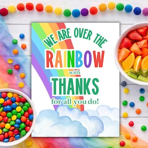 May include: A colorful print with the text "WE ARE OVER THE RAINBOW with THANKS for all you do!" features a rainbow and watercolor clouds. The image is surrounded by a bowl of fruit, a bowl of colorful candies, and a garland of pom-poms.