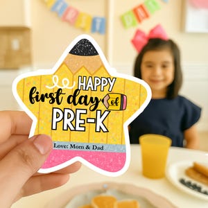 Puede incluir: Un letrero en forma de estrella con forma de lápiz con las palabras "Happy first day of PRE-K" en negro y amarillo. El letrero tiene una sección rosa y azul en la parte inferior con las palabras "Love: Mom & Dad".