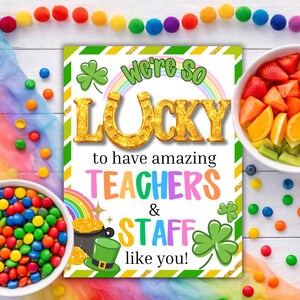 Op de afbeelding: Een St. Patrick's Day-bord met de tekst "We're So Lucky to have amazing TEACHERS & STAFF like you!" omringd door klavers, een regenboog en een pot met goud. Er zijn ook kleurrijke snoepjes en fruitschalen aanwezig.