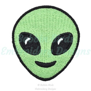 Puede incluir: Cara de extraterrestre verde con ojos negros y una boca sonriente. La cara del extraterrestre está bordada sobre un fondo blanco.