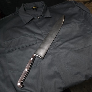 Pode incluir: Uma camisa de botões cinza-escura de manga comprida com colarinho e dois bolsos no peito, apresentando uma etiqueta preta com texto amarelo na gola. Uma faca de cozinha de metal grande com cabo de madeira e três rebites prateados.