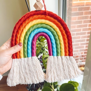 Puede incluir: Una pared colgante de hilo de colores arcoíris con una cuenta de madera en la parte superior. El arcoíris está hecho de múltiples hilos de hilo en diferentes colores, incluyendo rojo, naranja, amarillo, verde, azul, índigo y violeta. El hilo está atado en la parte superior con una borla blanca.
