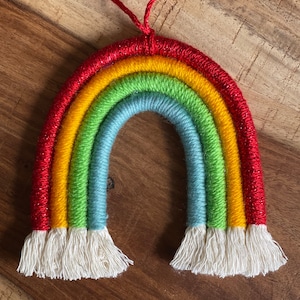 May include: A rainbow-coloured yarn wall hanging with a red, yellow, green, blue, and white colour scheme. The yarn is woven into a rainbow shape with a fringe at the bottom.