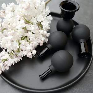 Peut inclure: Quatre bougeoirs noirs avec des bases rondes et un plateau noir. Les bougeoirs sont en verre et ont une finition mate. Un bouquet de fleurs de lilas blanches est sur le plateau.
