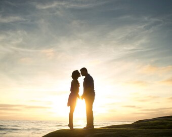 May include: Silhouette of a couple holding hands and about to kiss at sunset. The sky is a mix of blue, yellow, and orange. The couple is standing on a rock near the ocean. The sun is setting in the background.