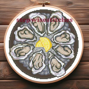 以下が含まれることがあります： レモンウェッジを添えたお皿に並べられたカキのクロスステッチ刺繍。刺繍は木のフープにセットされ、「stepwisestitches」の文字が特徴です。カキはグレーと白の色合いで、黄色のレモンスライスが付いています。