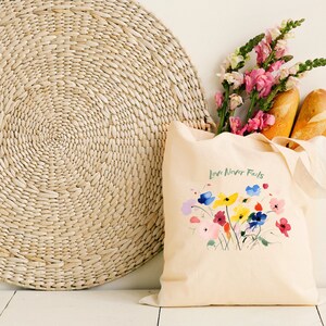 May include: A beige canvas tote bag with the text "Love Never Fails" printed on the front. The bag is decorated with a colourful floral design and has two long handles. The bag is sitting on a white surface next to a woven straw mat and a bouquet of pink and white flowers.