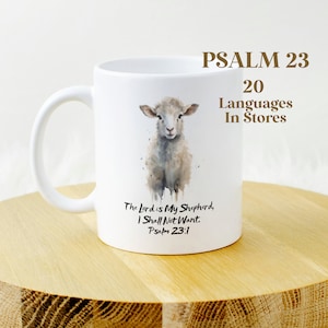 Może przedstawiać: Biały kubek ceramiczny z akwarelową ilustracją jagnięcia i tekstem "The Lord is My Shepherd, I Shall Not Want. Psalm 23:1". Kubek ma również tekst "PSALM 23 20 Languages In Stores" u góry.