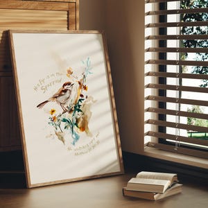 Puede incluir: Una pintura de acuarela de un gorrión posado en una rama con flores. El texto "His eye is on the sparrow" y "He watches over you" está escrito en cursiva en la impresión. La impresión está enmarcada en un marco de madera claro.