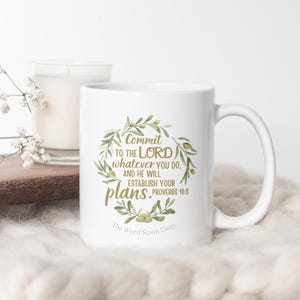 May include: White ceramic mug with a curved handle, featuring a gold and green olive branch wreath design. The text reads "Commit to the Lord whatever you do, and He will establish your plans. Proverbs 16:3." A candle and flowers are in the background.