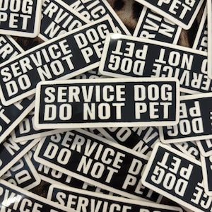 Op de afbeelding: Close-up van verschillende zwart-wit stickers met de tekst "Service Dog Do Not Pet".