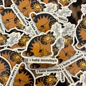 以下が含まれることがあります： 「月曜日は嫌い」というテキストと共に、タバコを吸っている漫画の猫が描かれたステッカーのコレクションです。