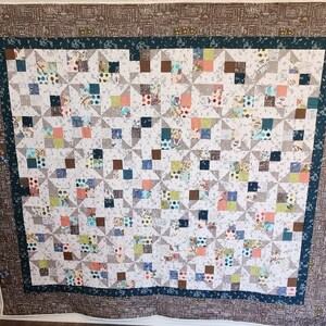 May include: A handmade quilt with a patchwork design. The quilt features a white background with a repeating pattern of squares in various colors, including blue, brown, pink, and green. The border is a dark brown fabric with a repeating pattern. The quilt is a cozy home decor item.
