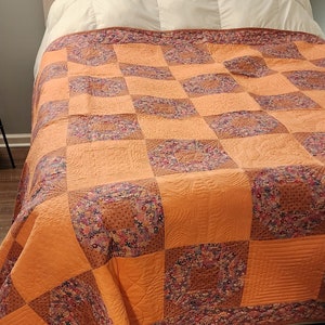 Può includere: Una trapunta fatta a mano con un design patchwork. La trapunta presenta grandi quadrati di tessuto arancione tinta unita e quadrati con motivi floreali nei toni del rosa, viola e marrone. La trapunta è su un letto con una testiera beige trapuntata.