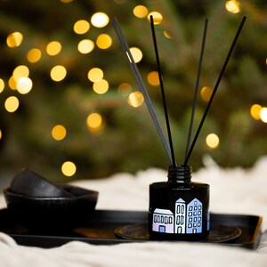 May include: A black glass reed diffuser bottle featuring a painted illustration of colorful houses in shades of pink and blue. The bottle contains several thin black reeds extending upwards and sits on a black rectangular tray next to a small black bowl.