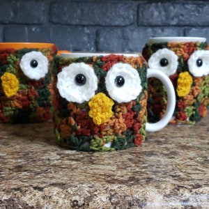 May include: Three crocheted owl mug cozies in shades of green, brown, and orange. Each cozy features a white owl face with black eyes and a yellow beak.
