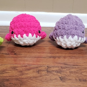 May include: Four handmade crochet shark plushies in a row. The sharks are in shades of green, pink, purple, and blue, each with white bellies and jagged white teeth. They have small black eyes and fins.