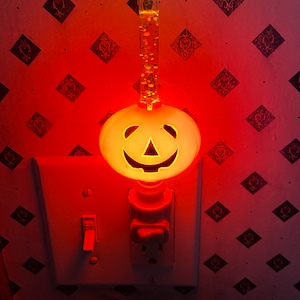 May include: A white pumpkin-shaped nightlight with a smiling jack-o'-lantern face is plugged into an electrical outlet. The nightlight is illuminated with a warm orange glow.