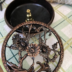 May include: A bronze-colored incense burner with a floral design on the lid. The lid features a circular pattern with flowers and leaves. The base is a dark brown color. A bundle of sage is in the background.