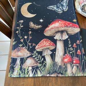 May include: A black table runner with a whimsical design featuring mushrooms, butterflies, and stars. The mushrooms are red and white with brown stems. The butterflies are brown and black. The stars are gold.