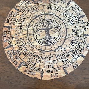 May include: A circular wooden calendar featuring a central tree illustration surrounded by concentric rings of text. The design includes months, moon names, seasonal festivals like Yule and Litha, and tree names with corresponding dates engraved in black.