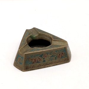 May include: A vintage brass ashtray in a triangular shape. The ashtray features a central circular depression and is decorated with teal and red floral patterns along the edges. The brass has a slightly aged appearance.