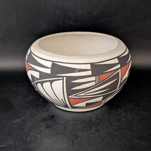 May include: A cream-colored, round ceramic bowl with a geometric design. The bowl features a pattern of black, white, and red shapes around the exterior. The interior of the bowl is plain cream. The bowl is likely used for decorative purposes.