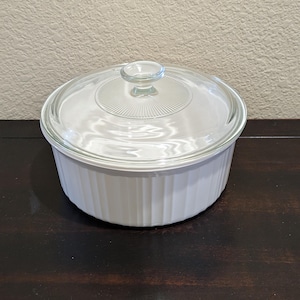 May include: A round white ceramic casserole dish with vertical fluted sides and a clear glass lid. The lid features a central knob handle and a radiating sunburst pattern etched into the glass around the center, providing a clear view inside.