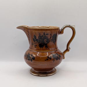 May include: A brown ceramic pitcher with a silver handle and black floral design.